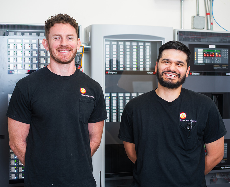 The owners of Real Protection Inc. standing in front of a fire alarm system