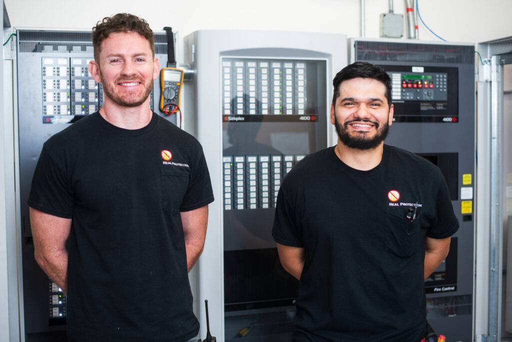 The owners of Real Protection Inc. standing in front of a fire alarm system