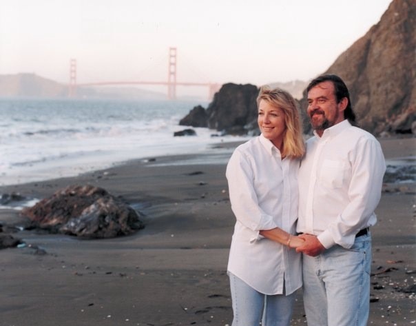 A blonde woman and a brunette man both wearing white button down shirts and light blue jeans on the beach, holding each other with the Golden gate bridge in the background.